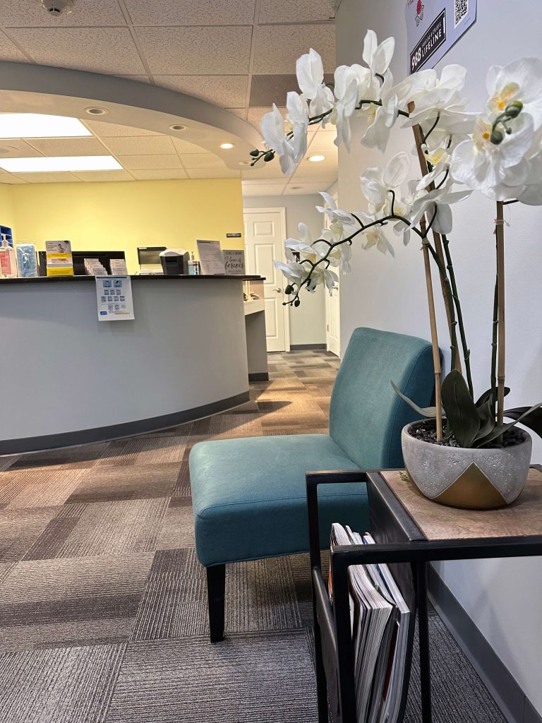 Image of reception area at NewVision Healthcare Group. The image contains a blue waiting room chair, the reception area and a white orchid in a neutral pot on a table next to the waiting room chair used for patients awaiting primary care or behavioral health services.